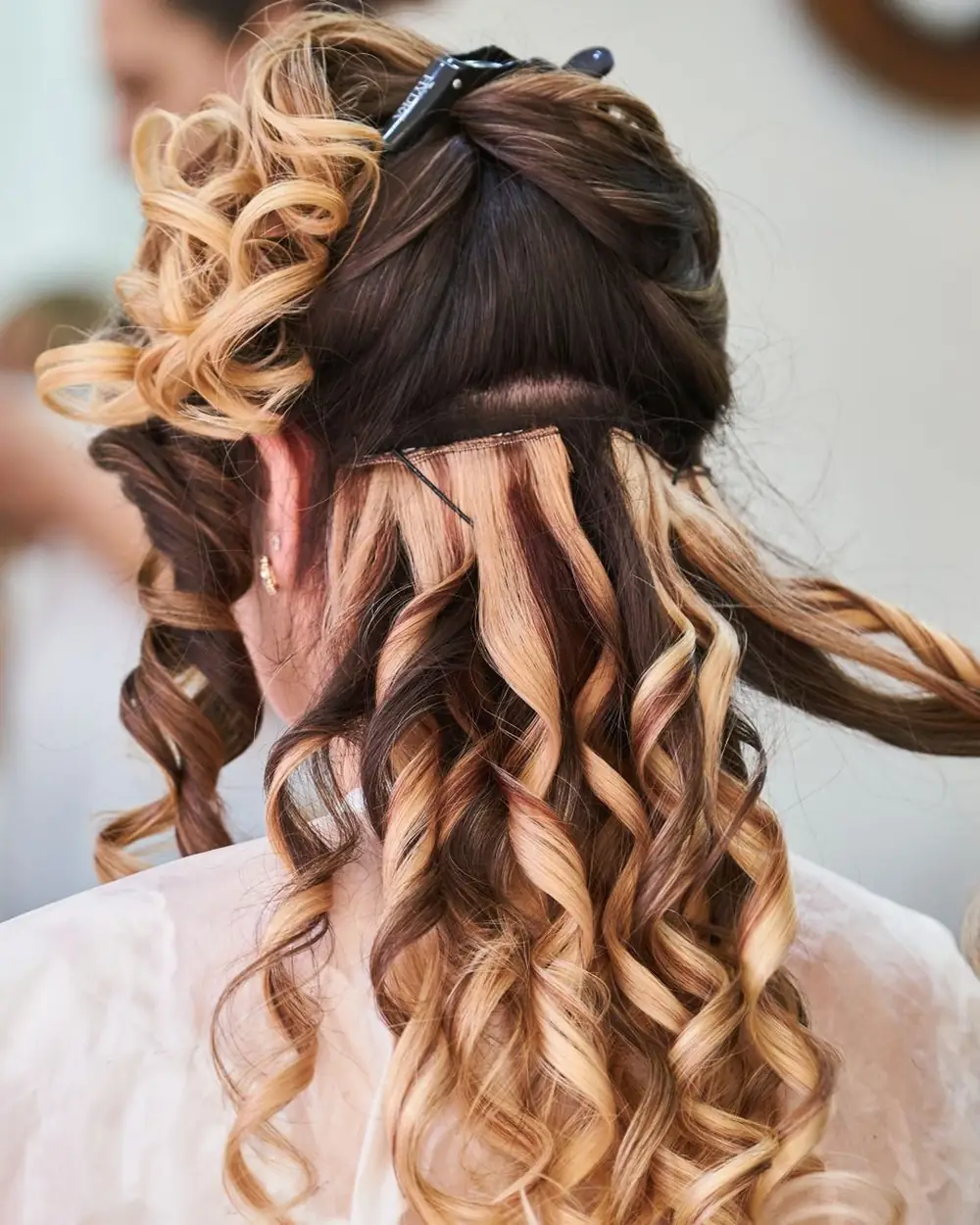 Back view of a stylist pinning long, glossy brown and blonde curls into a half-up event style